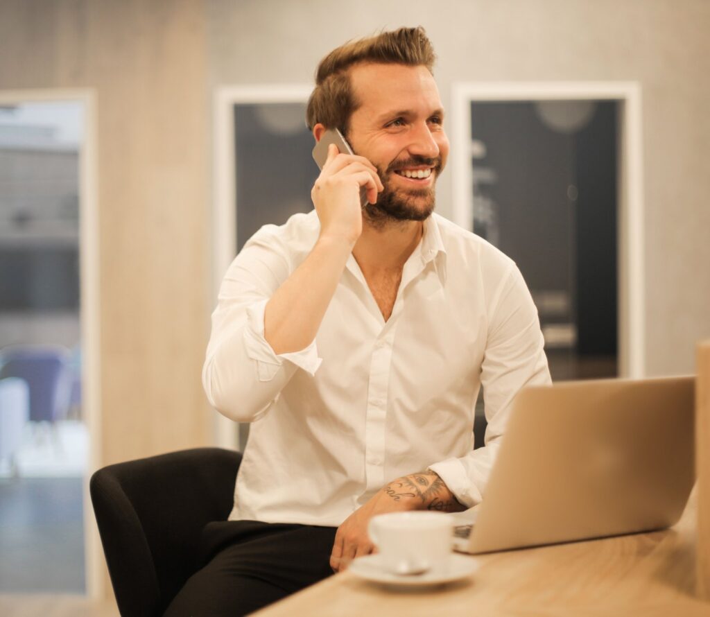 Mosolygó, barátságos kisvállalkozó telefonál a laptopja mellett az irodájában